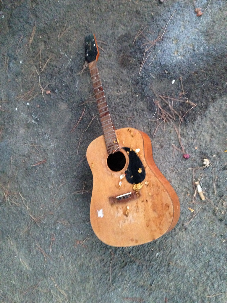 Damaged wooden acoustic guitar, laying on the ground or maybe concrete.
